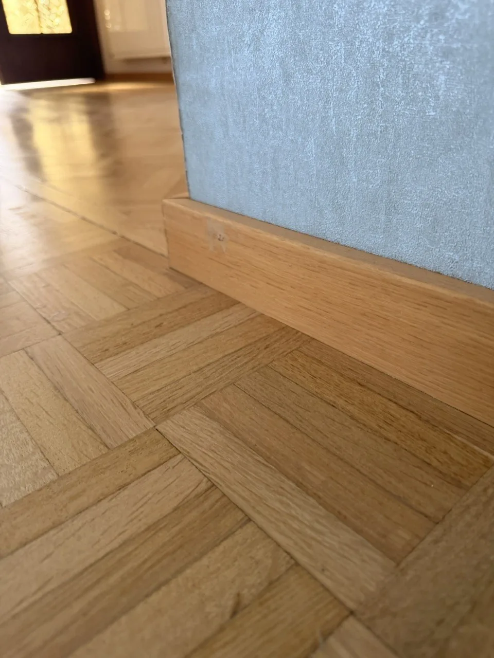 Détail de plinthes bois et de parquet mosaïque affleurant sous un papier peint clair