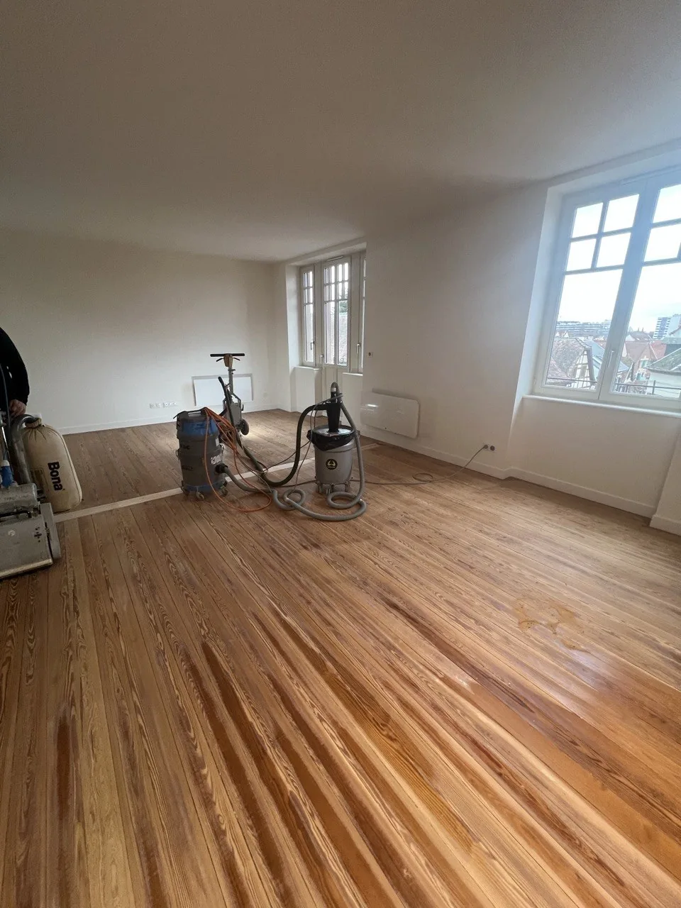 Ponceuse à bande et aspirateur de chantier raccordés sur un parquet mis à blanc dans une pièce vide