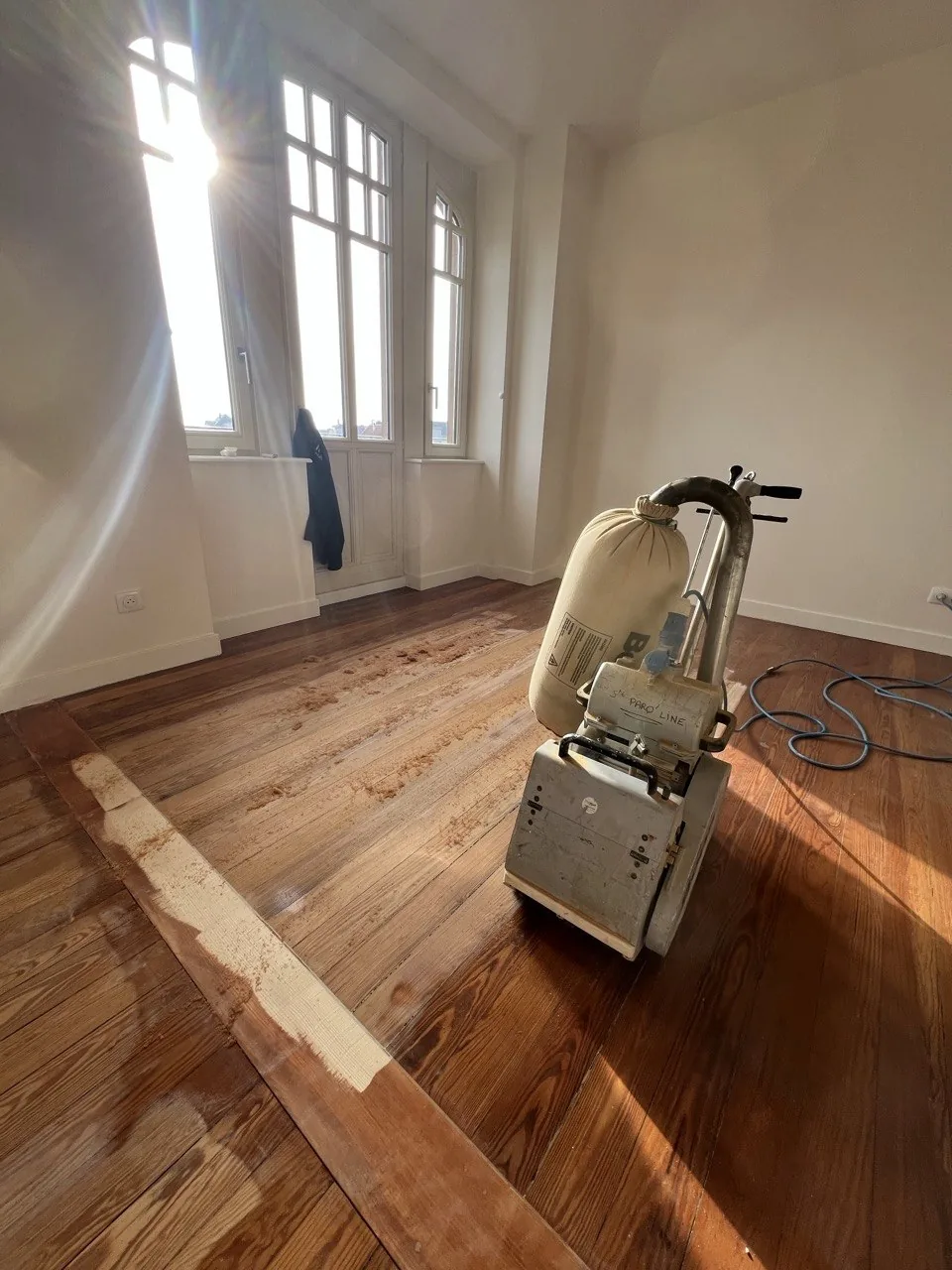 Ponceuse à bande de chantier marquée PARQ’LINE en train de mettre un parquet à nu, sciure sur le bois